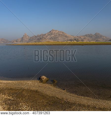 Landscape Of Jawai Dam With Water, Clear Blue Sky And Aravalli Mountain Ranges With Its Reflection I