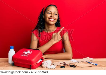 Young african american girl sitting on the table with first aid kit making cure smiling cheerful pointing with hand and finger up to the side 