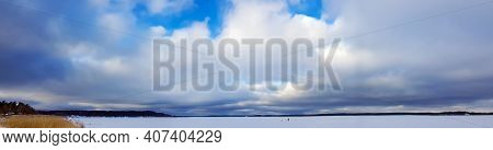 Panorama Of A Snowy Lake With A Lone Fisherman On The Background Of A Large Cumulus Clouds In Riga, 