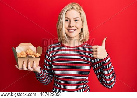 Young blonde woman eating fried chicken smiling happy and positive, thumb up doing excellent and approval sign 