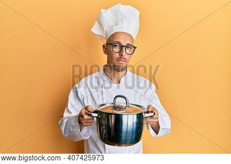 Bald man with beard wearing professional cook holding cooking pot relaxed with serious expression on face. simple and natural looking at the camera. 