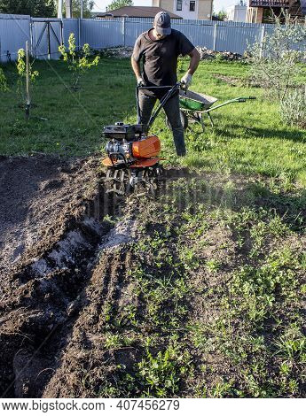 Male In The Spring Plows The Earth In Working Clothes With A Motoblock, Walk-behind Tractor