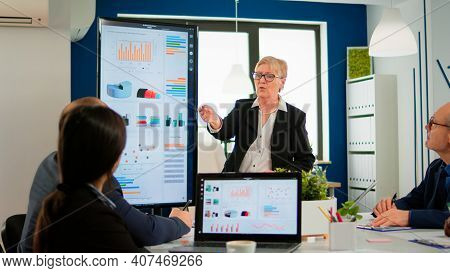 Elderly Project Manager Pointing At Desktop Presenting Statistical Data, Briefing Diverse Group Of E