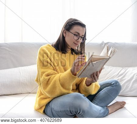 Stylish Brunette Girl In A Yellow Sweater And Glasses Reads A Book At Home On The Couch. The Concept
