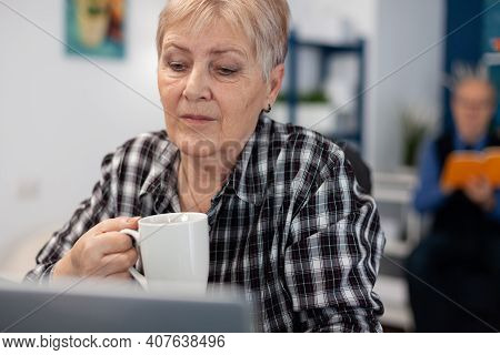 Senior Pensioner With Cup Of Coffee In Hands Reading Morning News. Elderly Woman In Home Living Room