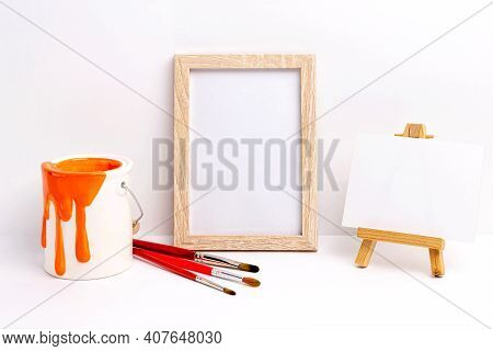 Mockup Of Blank Photo Frame And Canvas On Easel. Paint Bucket And Brushes. Concept Of Creativity, Ar