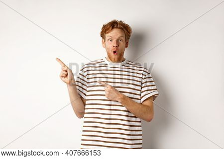 Amazed Man With Ginger Hair Checking Out Promo Offfer, Pointing Left At Logo And Smiling At Camera, 