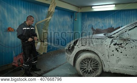 A Caucasian Man Works In A Car Wash Service. A Man In A Workers Uniform Sprays A Car With Detergent