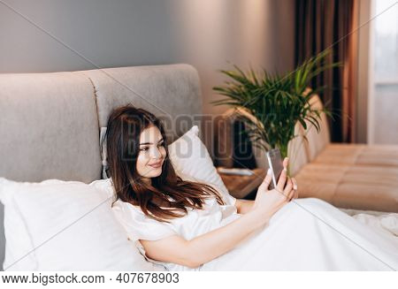 Young Woman Makes Selfie In Bed. The Woman Checks The Phone In The Morning. Brunette Scrolling The F