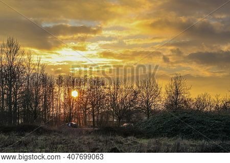Dutch Countryside With A Beautiful Sunset With The Sun Between The Branches Of The Bare Trees And Th