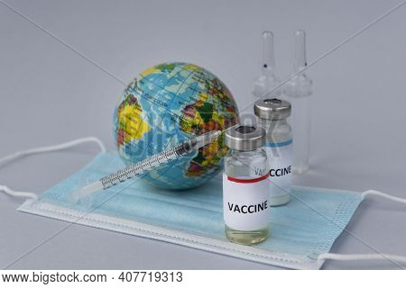 Two Ampoules With Vaccine And Inscription On Them, Syringe And Little Toy Globe On Medical Mask On L