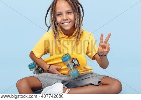 Cheerful Little Boy With African Dreads With Skateboard On The Blue Background Showing Peace Gesture