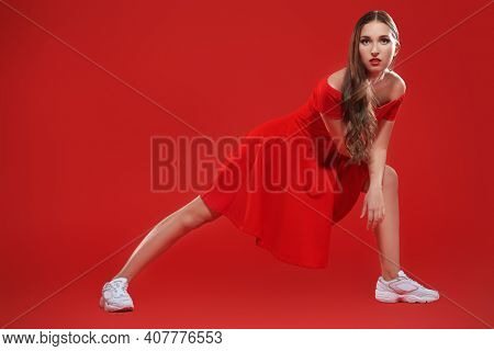 Full length portrait of a beautiful brunette girl posing at studio in red dress and white sneakers. Red background. Sport chic style. 