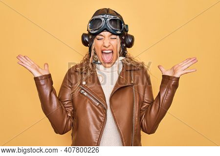 Young beautiful blonde aviator woman wearing vintage pilot helmet whit glasses and jacket celebrating mad and crazy for success with arms raised and closed eyes screaming excited. Winner concept