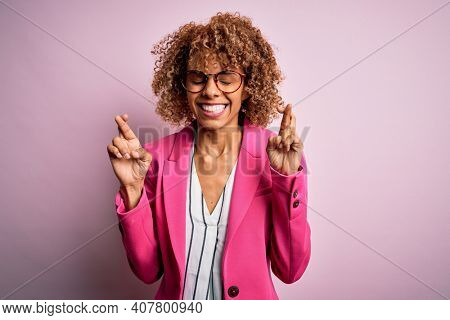 Young african american businesswoman wearing glasses standing over pink background gesturing finger crossed smiling with hope and eyes closed. Luck and superstitious concept.