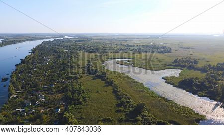 Drone Fly Over Waving River Of Blue Color Surrounded By Local Village With Various Buildings And Wet