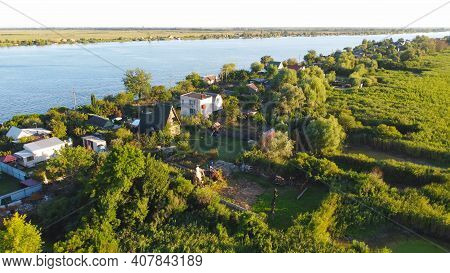Drone Fly Over Waving River Of Blue Color Surrounded By Local Village With Various Buildings And Wet
