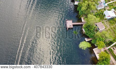 Drone Fly Over Waving River Of Blue Color Surrounded By Local Village With Various Buildings And Wet