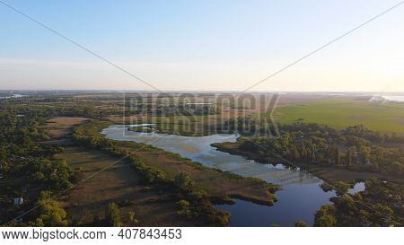 Drone Fly Over Waving River Of Blue Color Surrounded By Local Village With Various Buildings And Wet