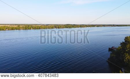 Drone Fly Over Waving River Of Blue Color Surrounded By Local Village With Various Buildings And Wet
