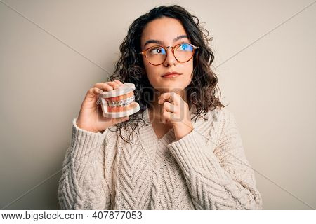 Young beautiful woman with curly hair holding plastic teeth with dental baces serious face thinking about question, very confused idea