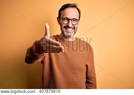 Middle age hoary man wearing brown sweater and glasses over isolated yellow background smiling friendly offering handshake as greeting and welcoming. Successful business.