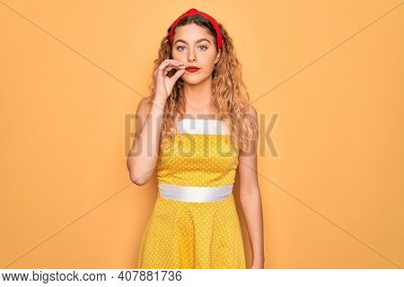 Beautiful blonde pin-up woman with blue eyes wearing diadem standing over yellow background mouth and lips shut as zip with fingers. Secret and silent, taboo talking