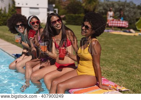 Diverse group of female friends drinking drinks sitting at the poolside. Hanging out and relaxing outdoors in summer.
