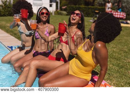 Diverse group of female friends drinking drinks sitting at the poolside. Hanging out and relaxing outdoors in summer.