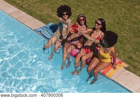 Diverse group of female friends drinking drinks sitting at the poolside. Hanging out and relaxing outdoors in summer.