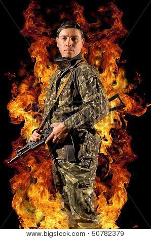 Soldier stands with a gun in his hand and safety glasses in a burning fire