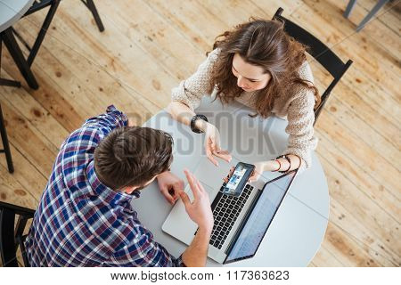 Top view of young happy couple sitting at round table in cafe and looking at screen of mobile phone and lapop