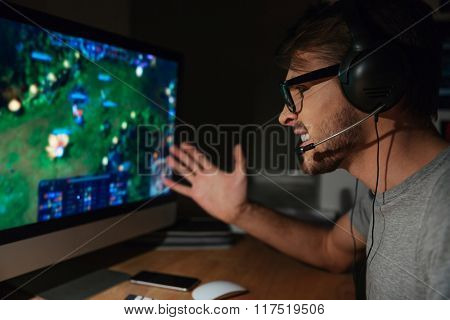 Angry young gamer in glasses playing game on computer using headphones 