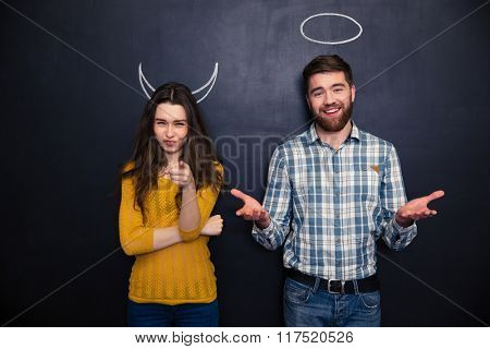 Beautiful young couple standing over blackboard background with drawn hornes and halo and pointing on you