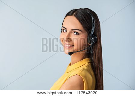 Photo of beautiful young call center operator standing near gray background. Woman with headphones looking at camera and smiling
