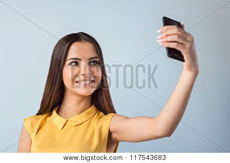 Photo of beautiful young business woman standing near gray background. Woman with yellow shirt making selfie with mobile phone and smiling