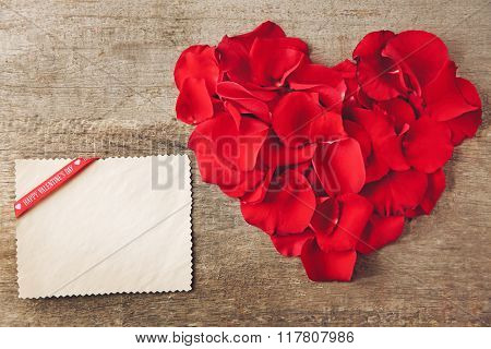 Heart made of rose petals with blank card on wooden background