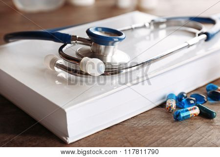 Stethoscope with open book and pills on wooden background. Medical literature concept
