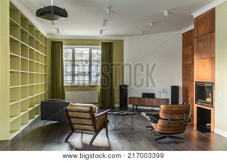 Room in a modern style with light walls and a parquet on the floor. There are shelves, stylish armchairs, sofa, round table, fireplace with brown chimney, wooden rack, speakers, lamps, projector.