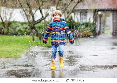 Little kid boy wearing yellow rain boots and walking during sleet, rain and snow on cold day. Child in colorful fashion casual clothes jumping in a puddle. Having fun outdoors.
