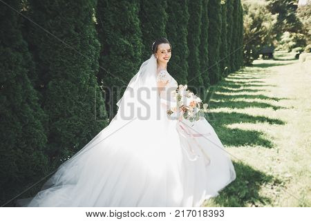 Beautiful bride in elegant white dress holding bouquet posing in park.