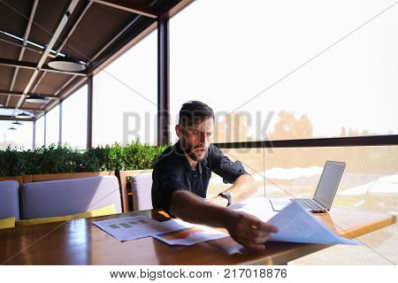 Placeman sorting papers on desk near laptop. Strong male hands with watch near window. Concept of stopping document clutter on workplace.