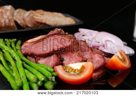 business lunch fresh roast beef meat slices on black plate wotj cutlery asparagus boiled broccoli rye bun on wooden table