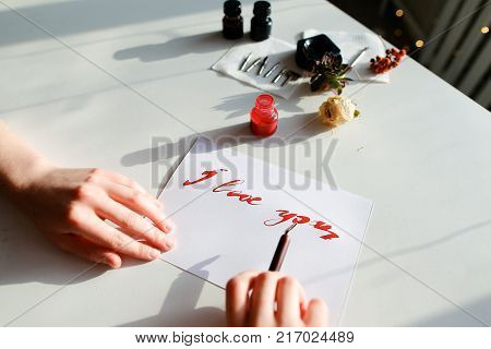 Female master of calligraphy and lettering creates red ink and pen fountain with wonderful love valentines with text I love you on day of all lovers on light table with accessories for lettering, small jars with colorful mascara and decorations in art stu