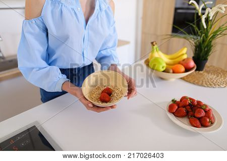 Slender, sweet woman cooks delicious breakfast of oatmeal and ripe red strawberry, with smile on face, takes plate in hands and brings it to camera lens, standing in stylish and light kitchen with white cabinets, household appliances and variety of utensi