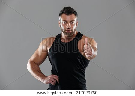 Portrait of a serious strong male bodybuilder standing and showing thumbs up gesture isolated over gray background