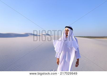 Cheerful Muslim guy walks along white sand of wide desert on warm summer evening. Arab young man in national clothes dances on spot and laughs giggly, adjusting hands with harness in wind. Swarthy, handsome Muslim with short dark hair dressed in kandura, 