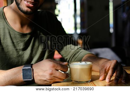 Close-up shooting, man sitting in cafe and drinking hot coffee with milk. Boy dressed in khaki pullover. Attractive mullato has dimples, beard, full lips and black hair. Concept of modern places for young people cheap prices.