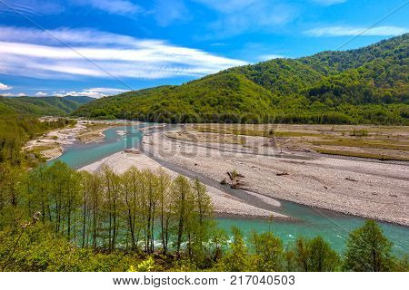 The azure mountain river flows around the stones between the forest and the mountains against the background of a bright blue sky in spring or summer. Shahe river, Bolshoy Kimchay, Sochi, Russia.