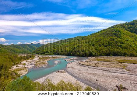 The azure mountain river flows around the stones between the forest and the mountains against the background of a bright blue sky in spring or summer. Shahe river, Bolshoy Kimchay, Sochi, Russia.